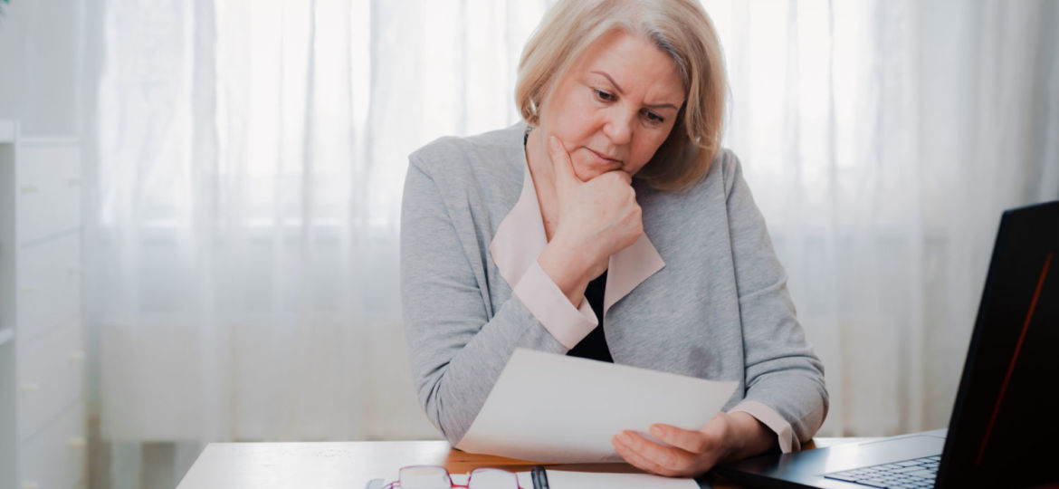 older Senior woman struggles at the computer with paper documents, puzzled by the business decision